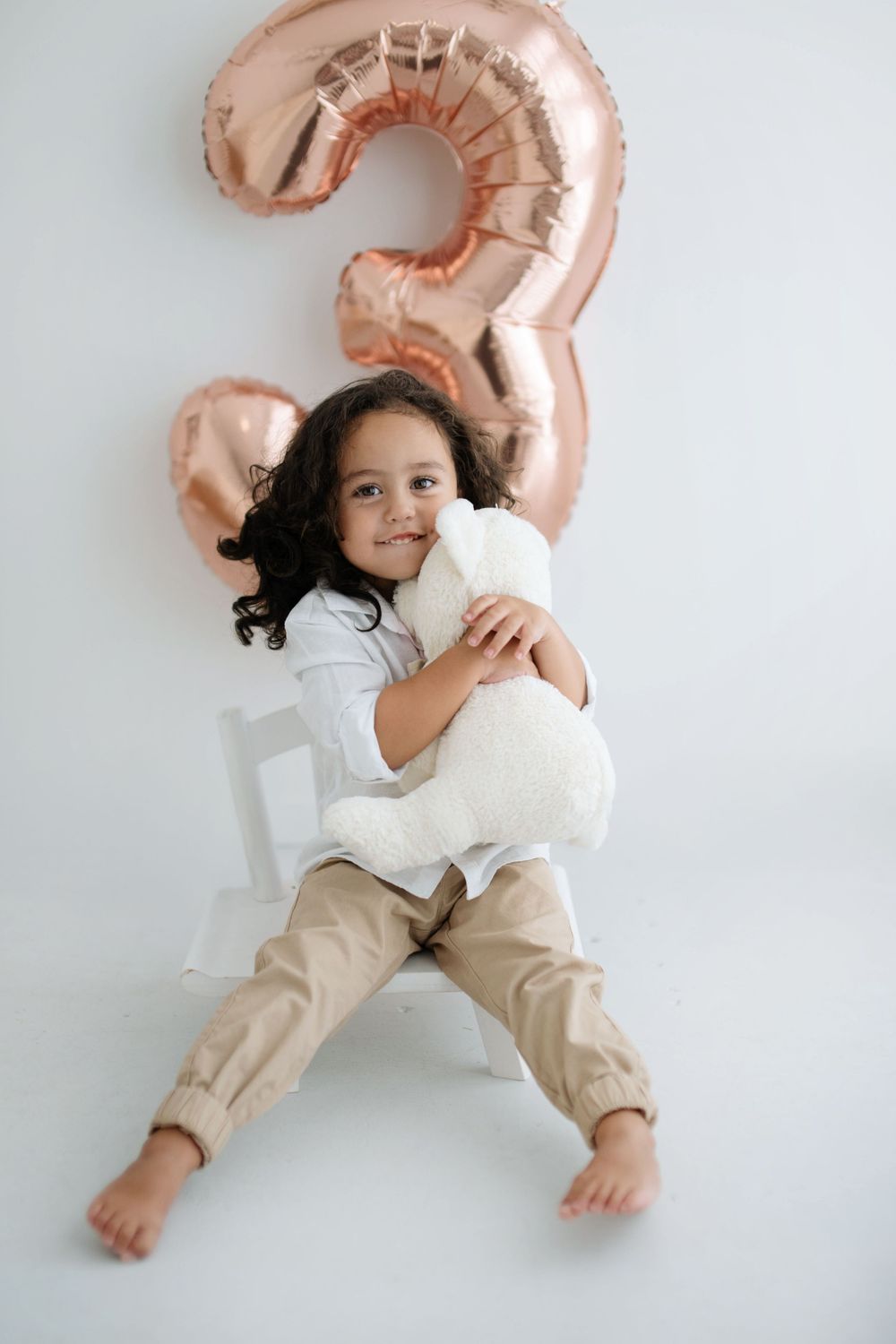 A young girl hugs a white teddy bear while sitting in front of a rose gold number 3 balloon on her birthday.