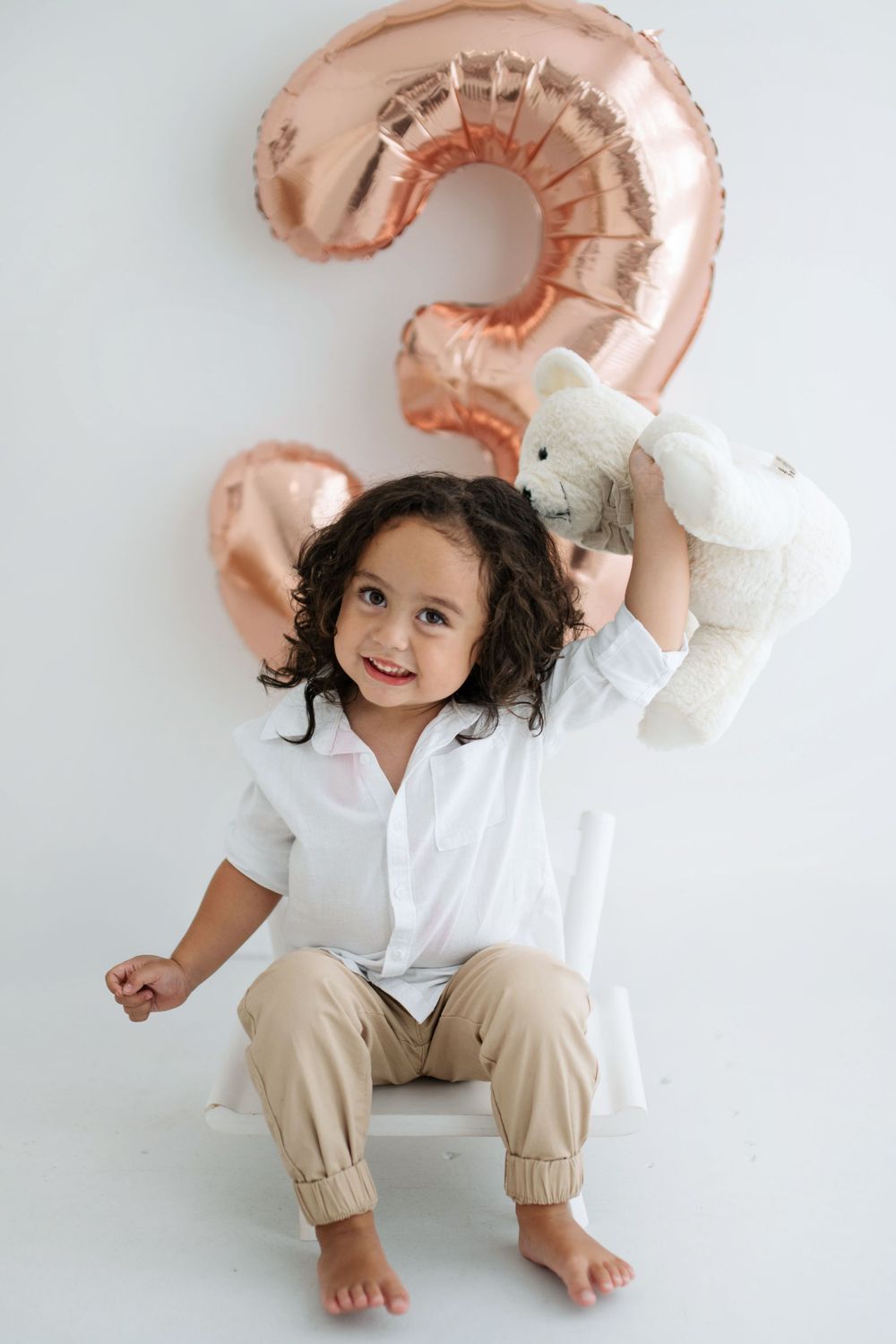 A happy toddler holds a white teddy bear in front of a rose gold number 3 birthday balloon.