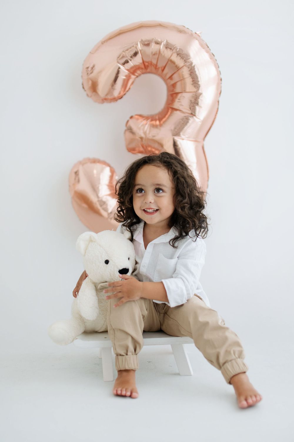 A smiling child sits with a white teddy bear in front of a rose gold number 3 balloon.