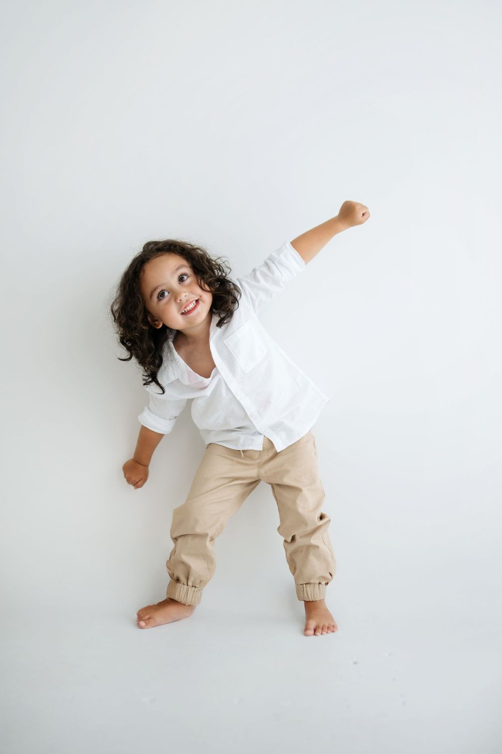A joyful young child with curly hair poses in a white shirt and tan pants, raising one fist up like a superhero.