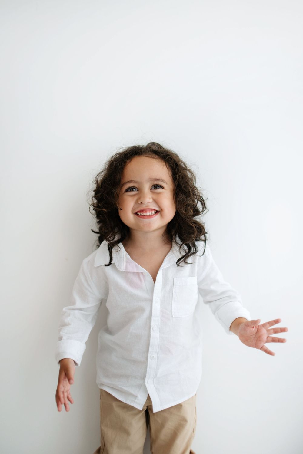 A happy curly-haired child in a white button-up shirt smiles with arms outstretched against white wall.