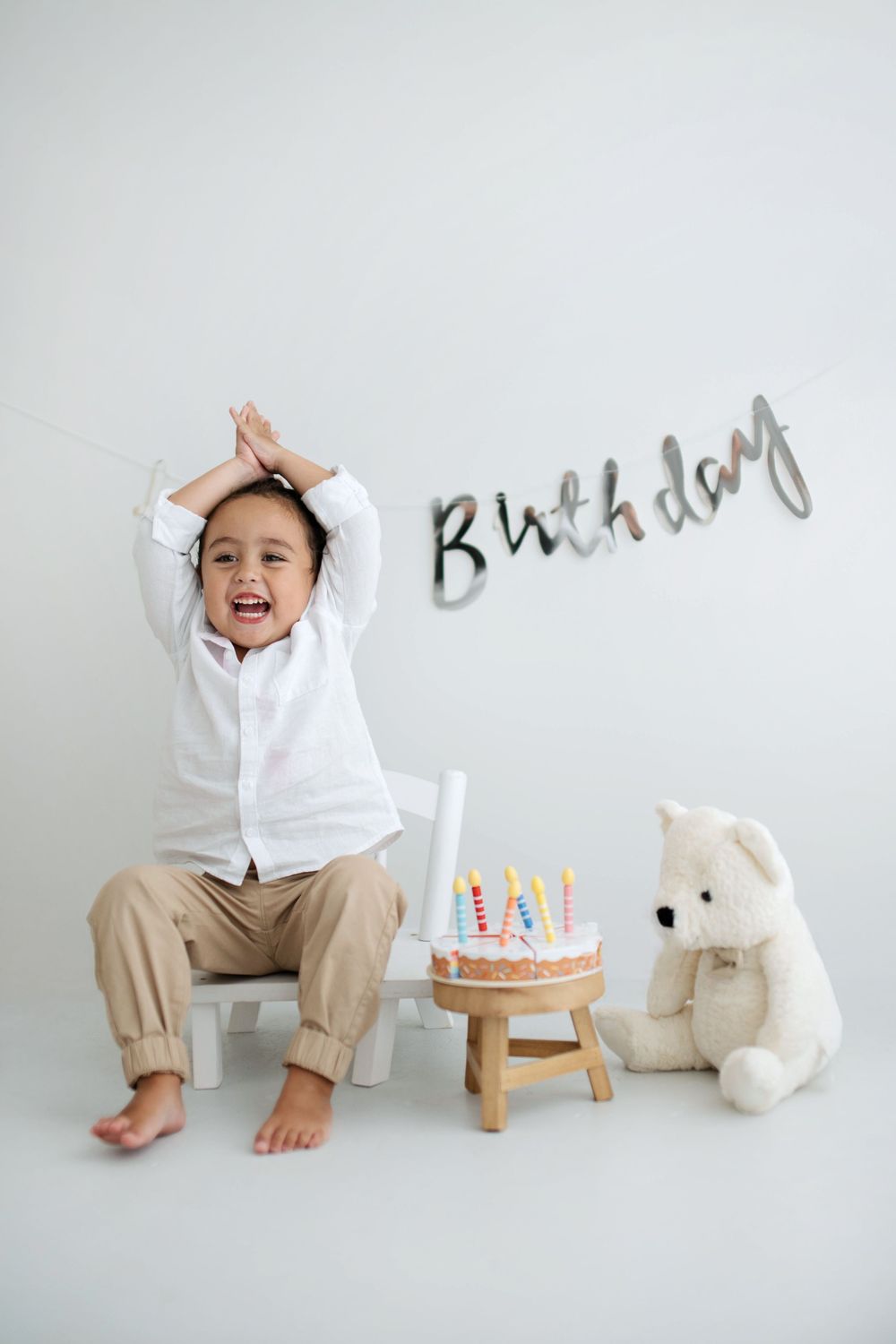 A joyful young boy celebrating his birthday with a cake and teddy bear, wearing a white shirt and khaki pants.