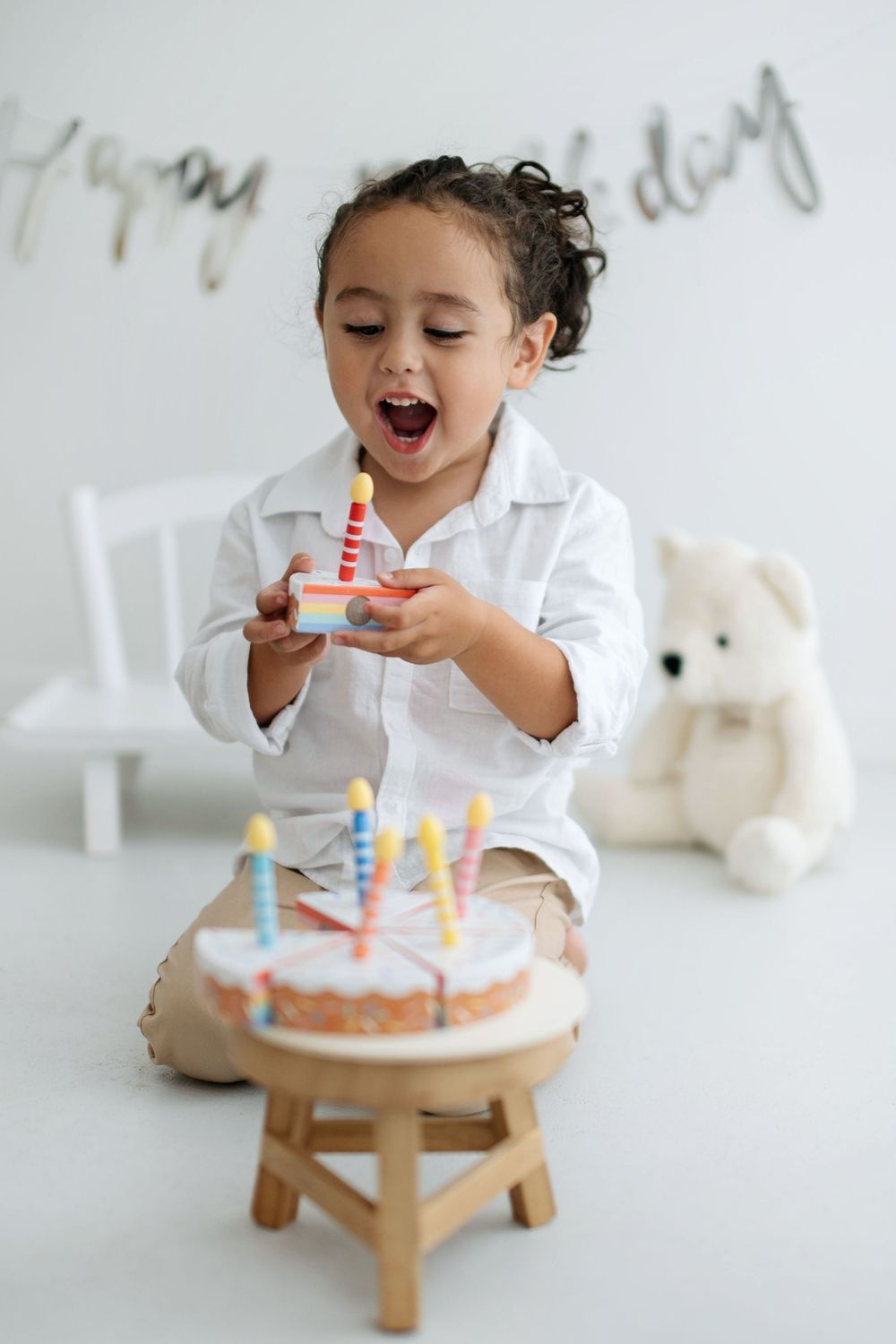 A joyful toddler in a white shirt celebrates their birthday with a colorful cake and candles in a bright white room.