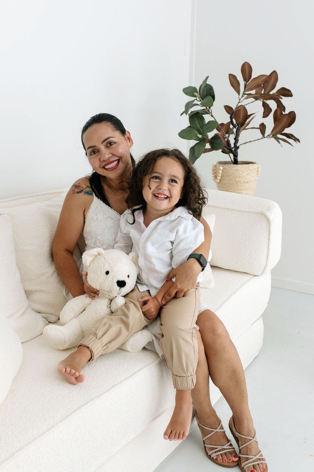 A smiling woman and young child sit together on a white sofa, holding a teddy bear, with a potted plant nearby.