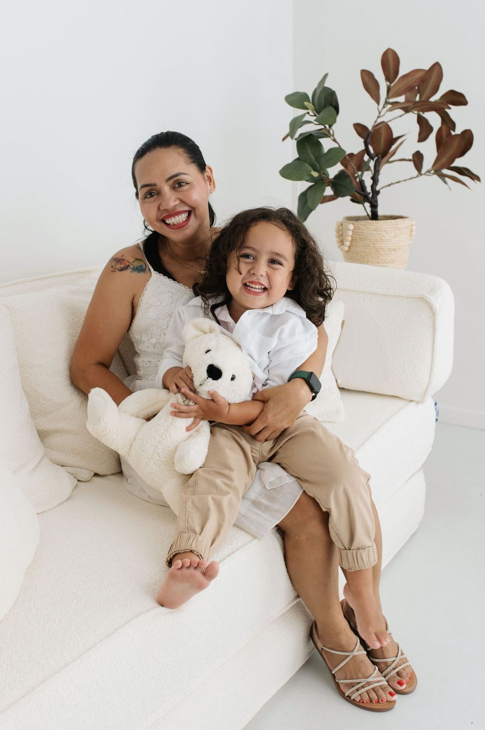 A smiling woman and young child sit together on a white sofa, the child holding a white teddy bear.