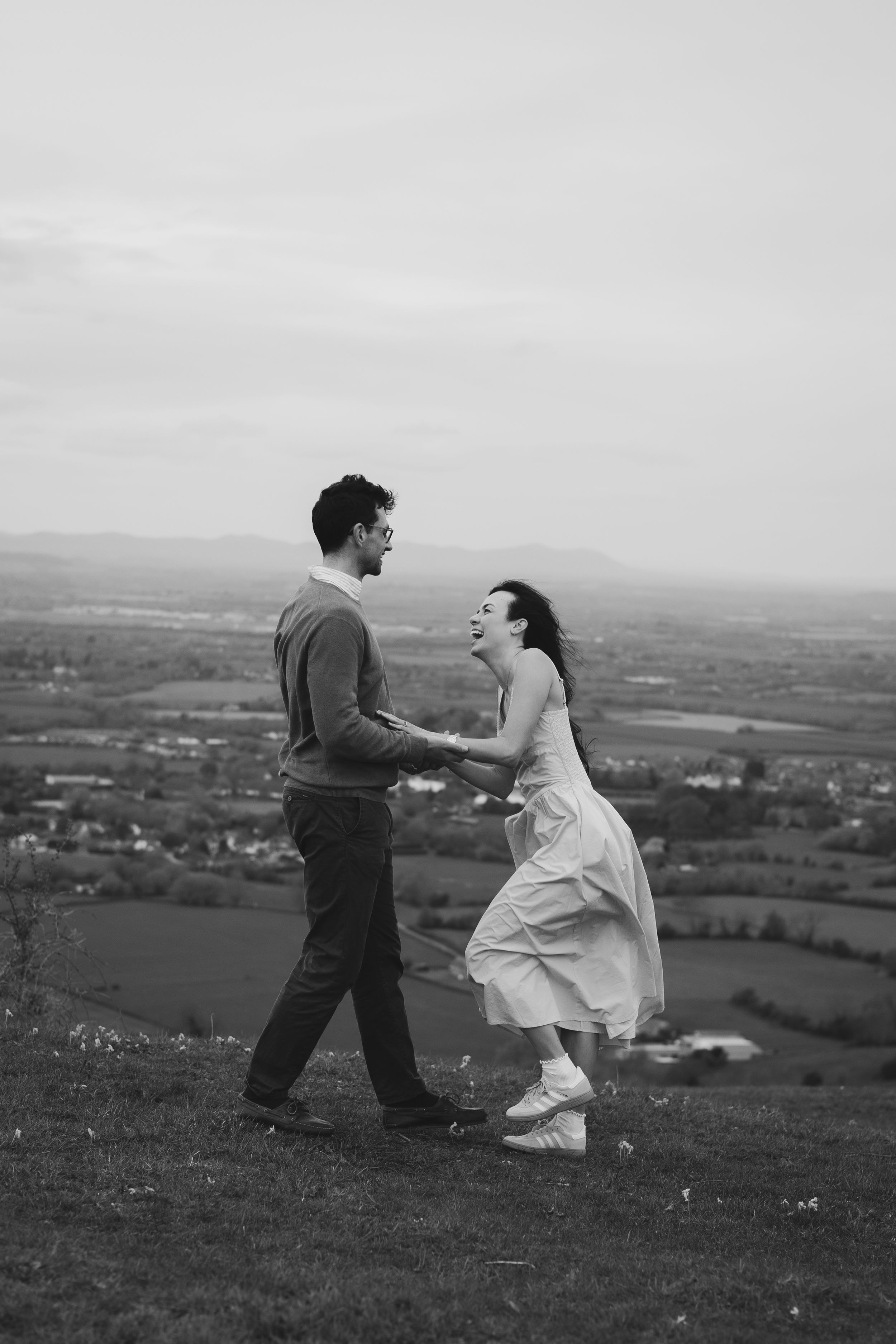 Black and white photo of a laughing bride in white dress and groom holding hands on a hilltop with scenic countryside views.