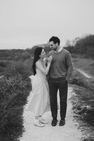 Black and white photo of a couple smiling at each other on a scenic outdoor path, woman in white dress, man in sweater.
