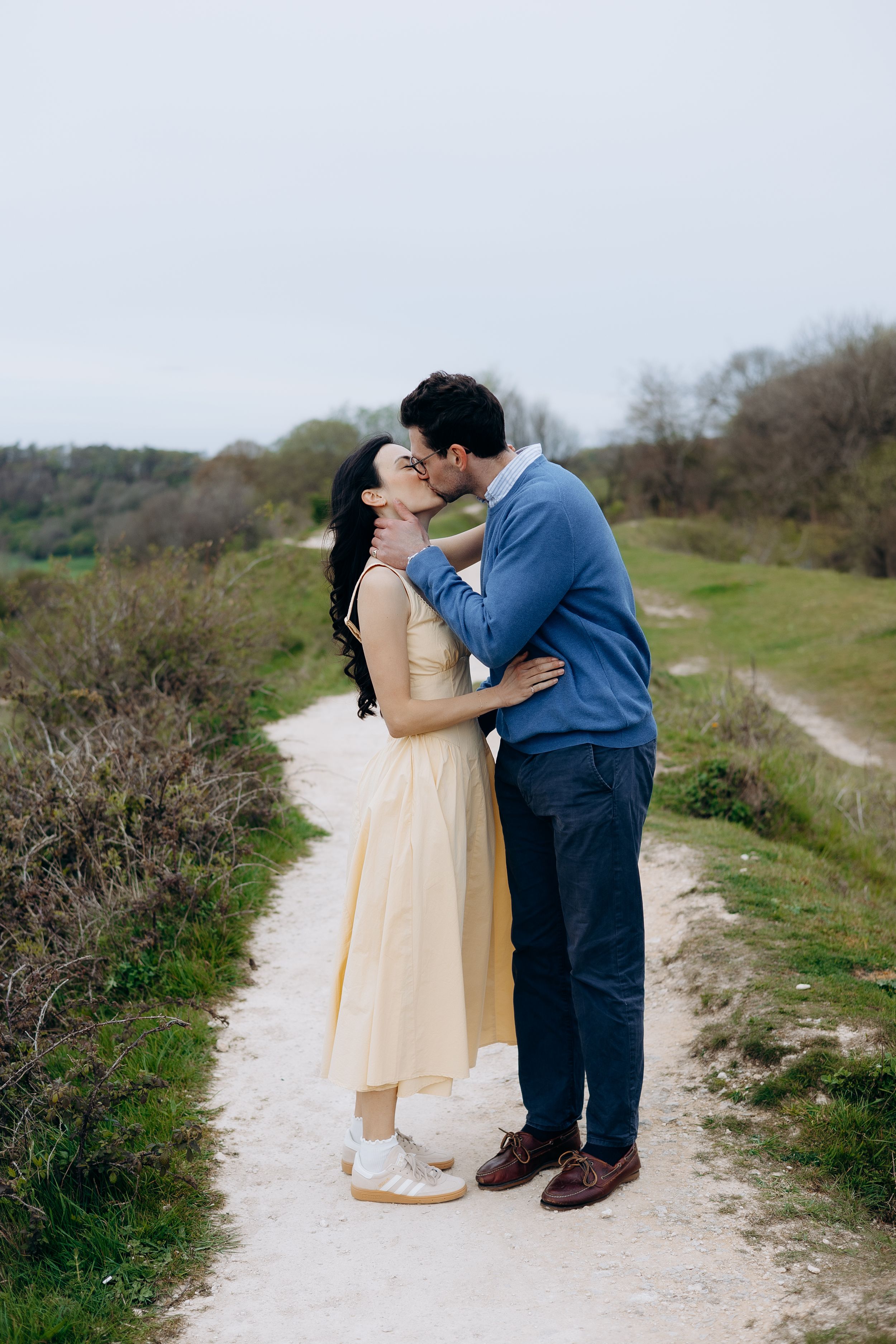Couple sharing a romantic kiss on a scenic dirt path surrounded by lush greenery, woman in yellow dress, man in blue jacket.