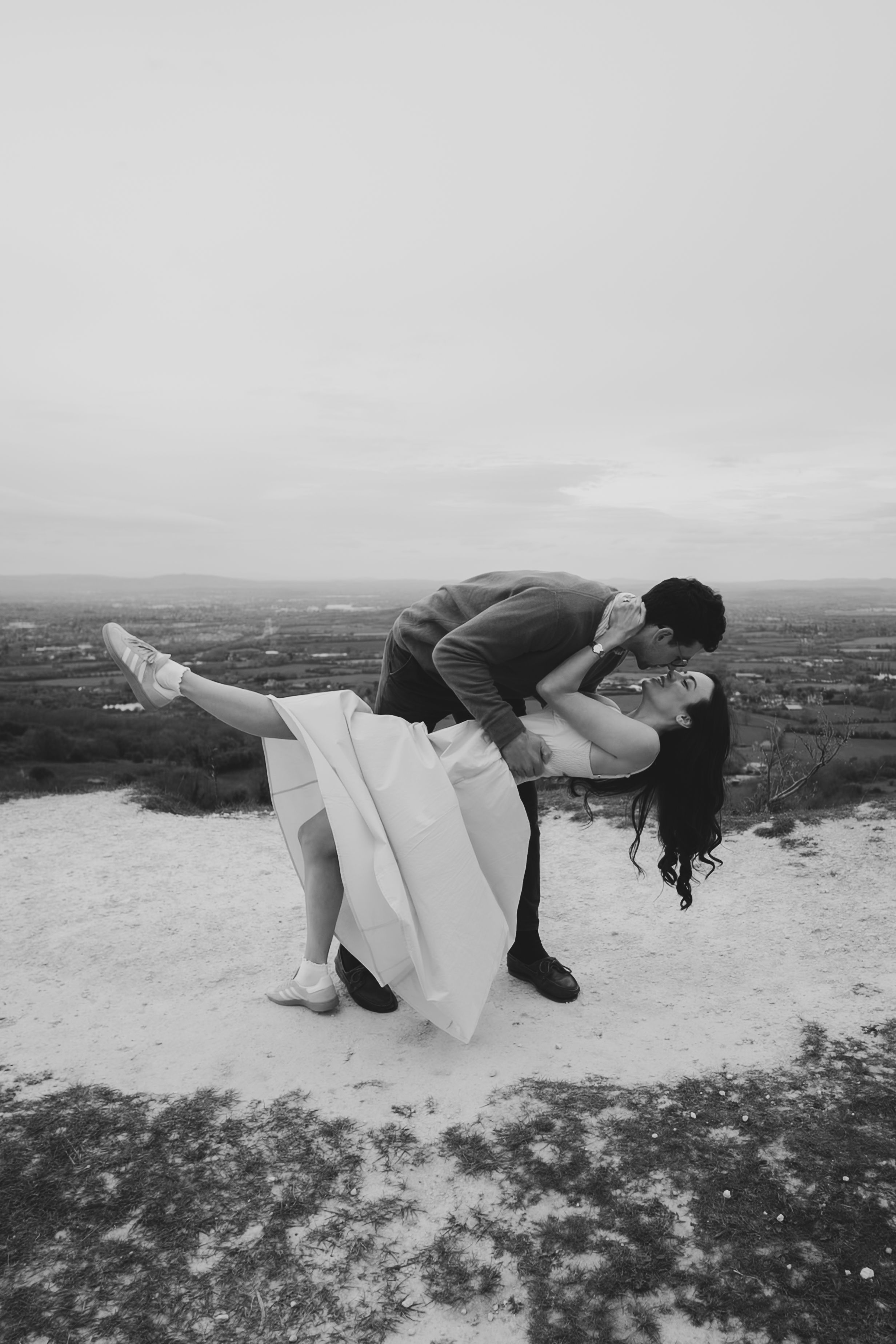 Black and white photo of a groom dipping his bride for a romantic kiss on a hilltop with scenic landscape views.
