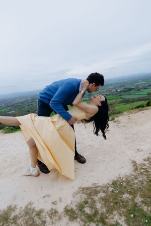 A couple shares a romantic dip kiss on a hilltop with scenic green countryside views in the background.