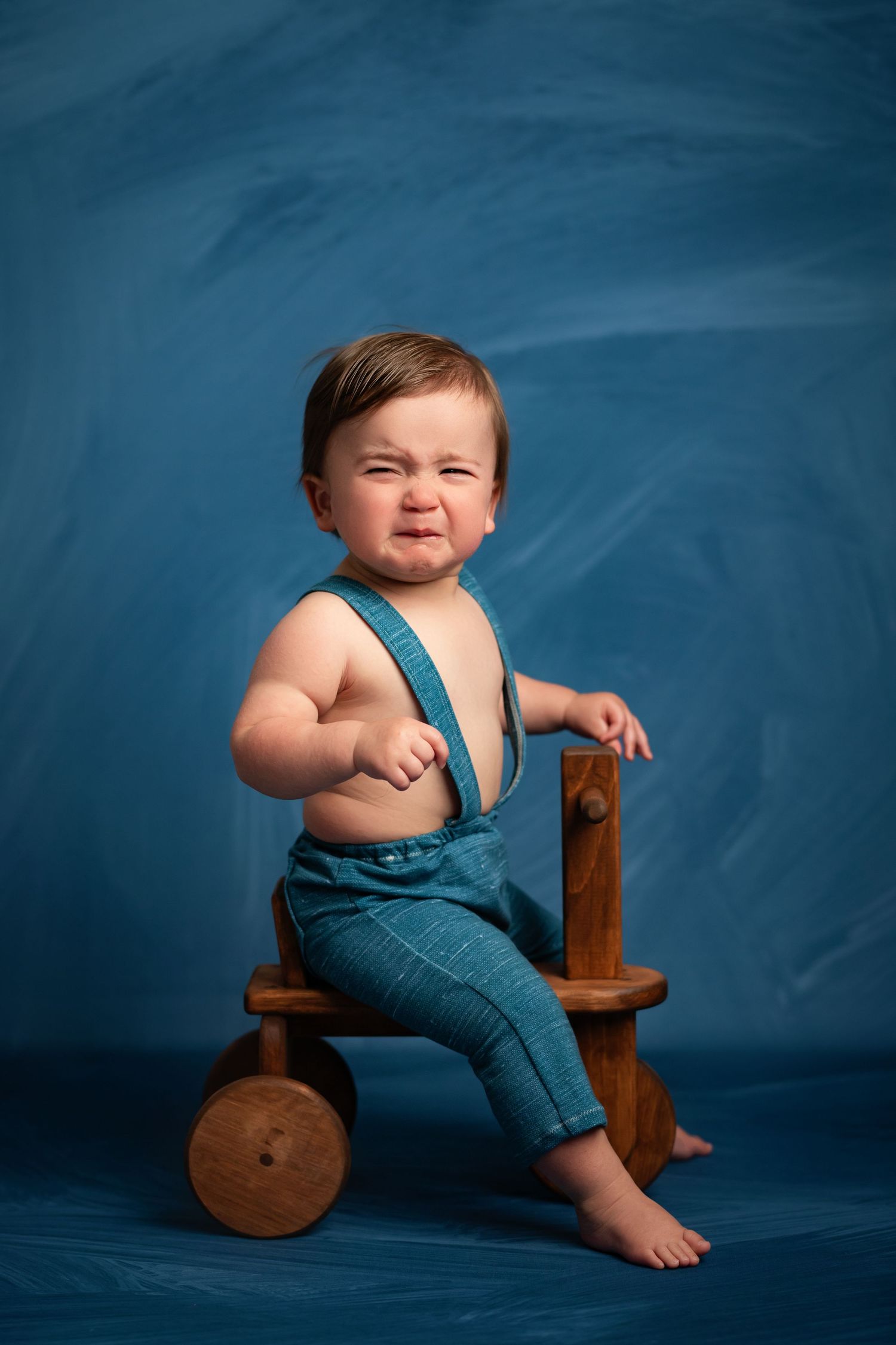 Cute baby boy in blue suspenders making a funny face while sitting on a wooden toy cart against blue background.