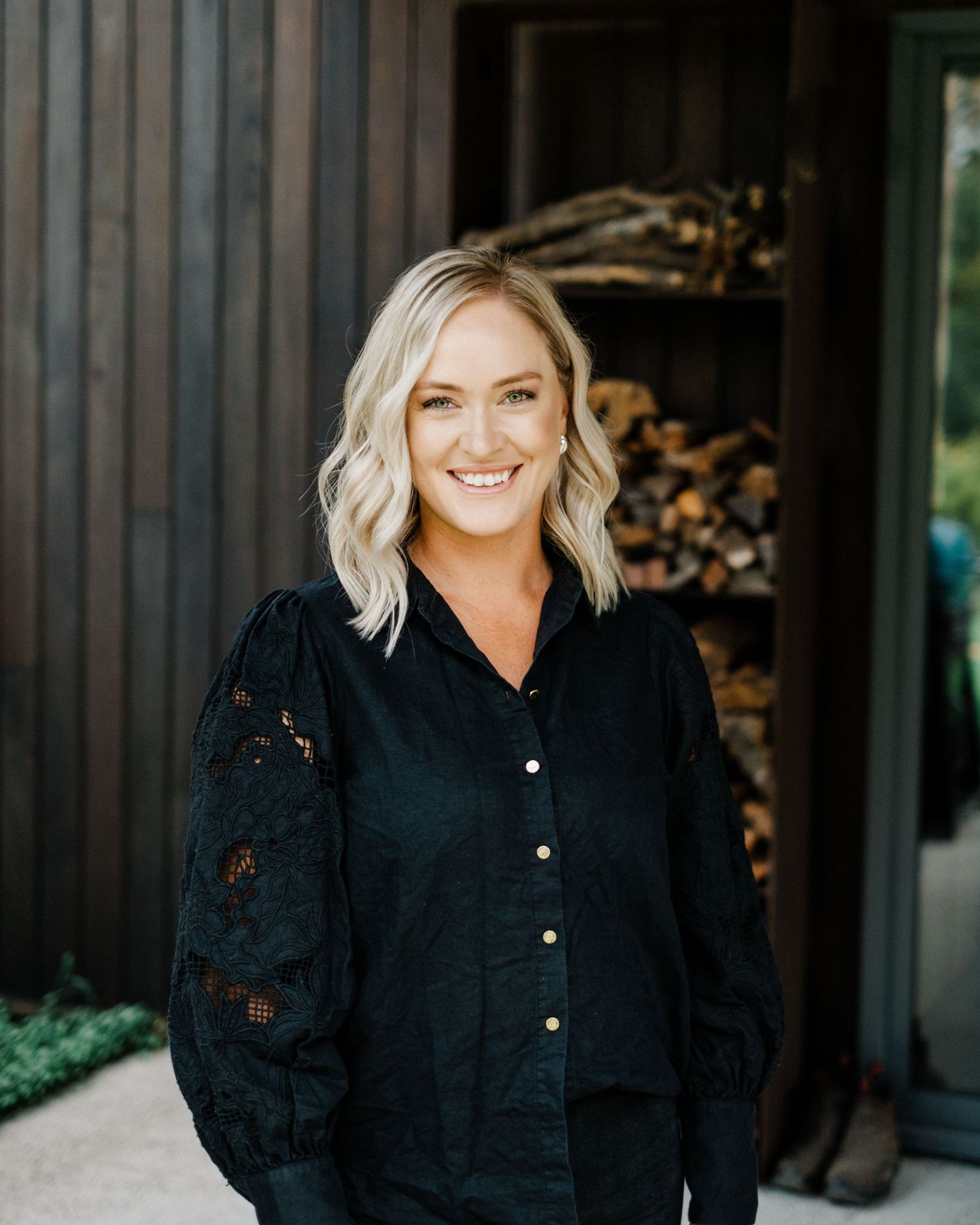 Smiling blonde woman in black lace blouse standing outdoors near dark wood paneling and stacked firewood.