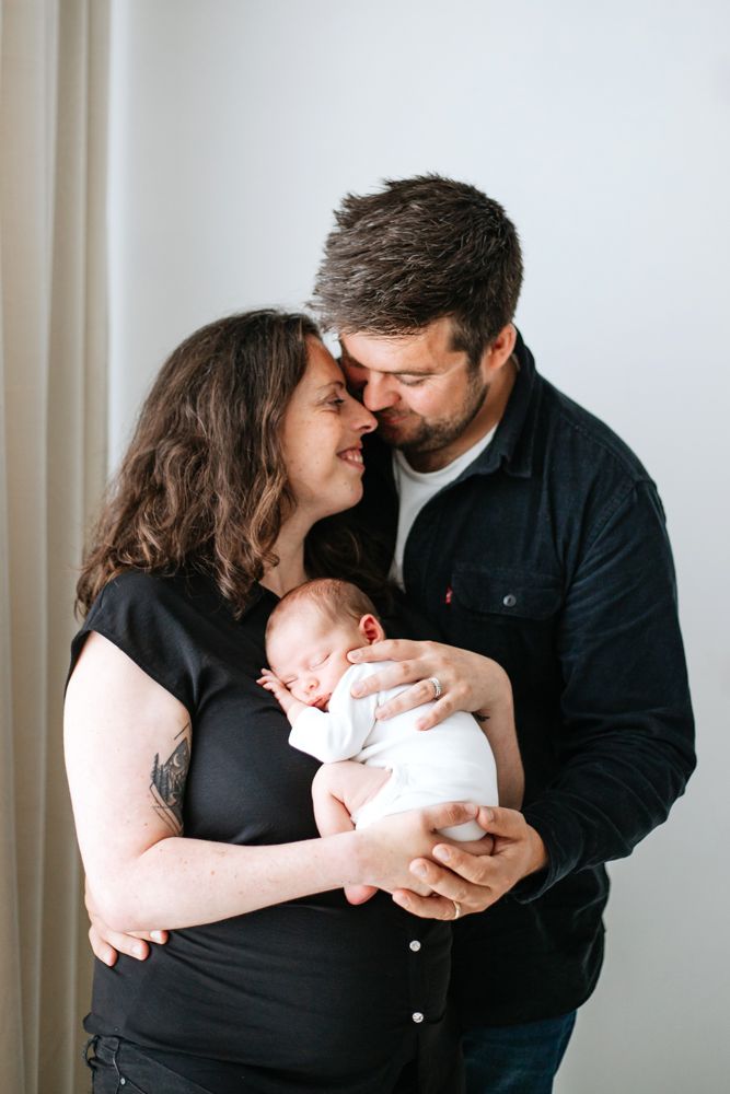 A couple tenderly nuzzles while holding their swaddled newborn baby, capturing a precious family portrait moment.