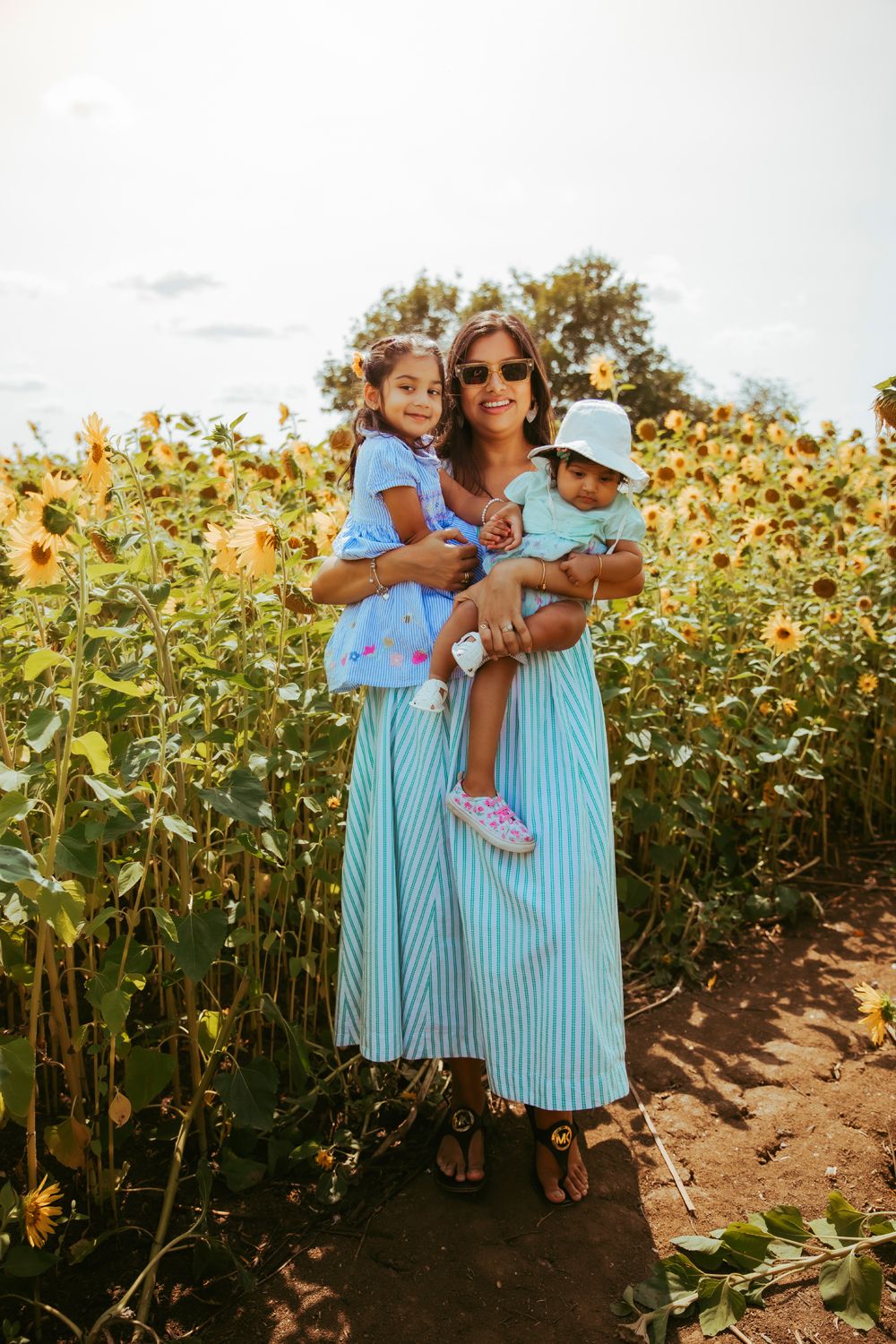 Woman in blue striped dress holding two young girls in a sunflower field on a sunny day. At a photoshoot in London