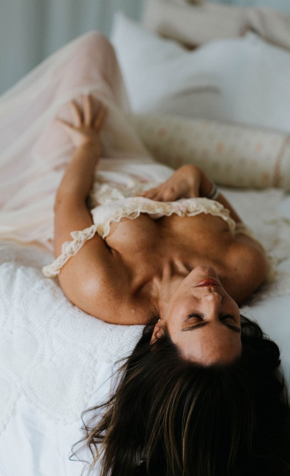 Brunette woman lying on white bed wearing cream lace lingerie in sensual boudoir portrait.