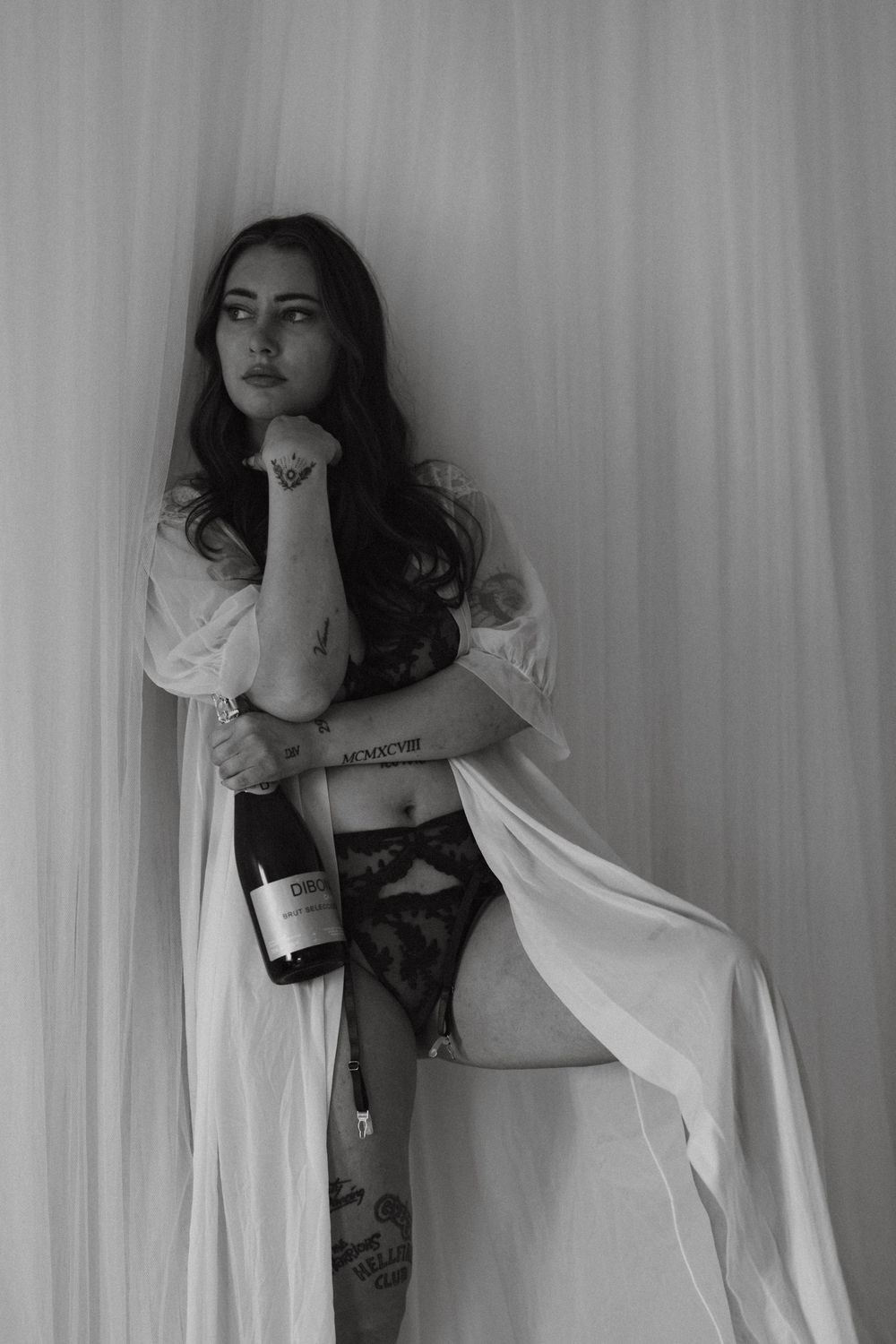 Black and white boudoir photo of tattooed woman in lingerie holding champagne bottle by sheer curtains.