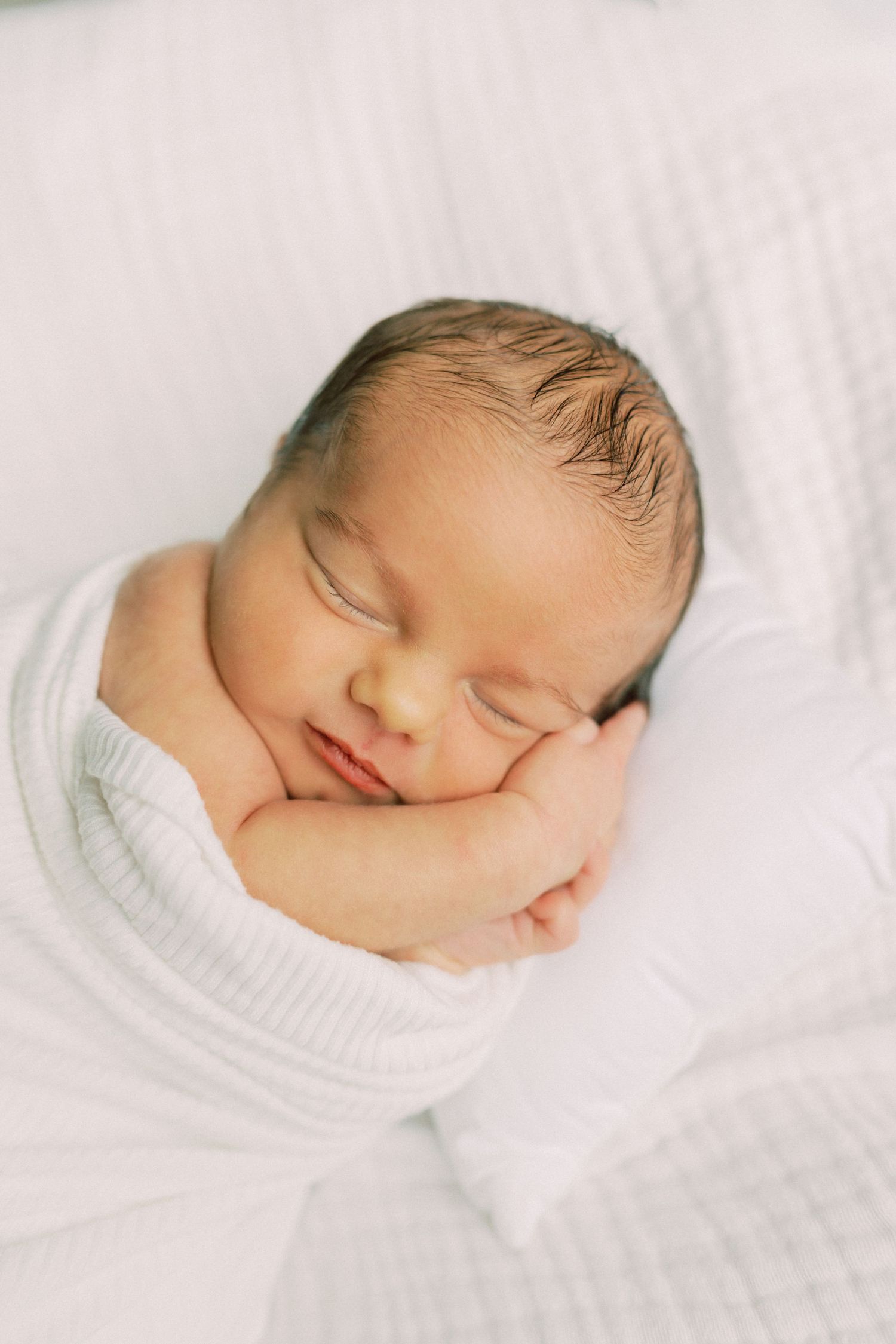 Peaceful newborn baby sleeping soundly wrapped in a soft white swaddle blanket with tiny hands tucked under cheek.