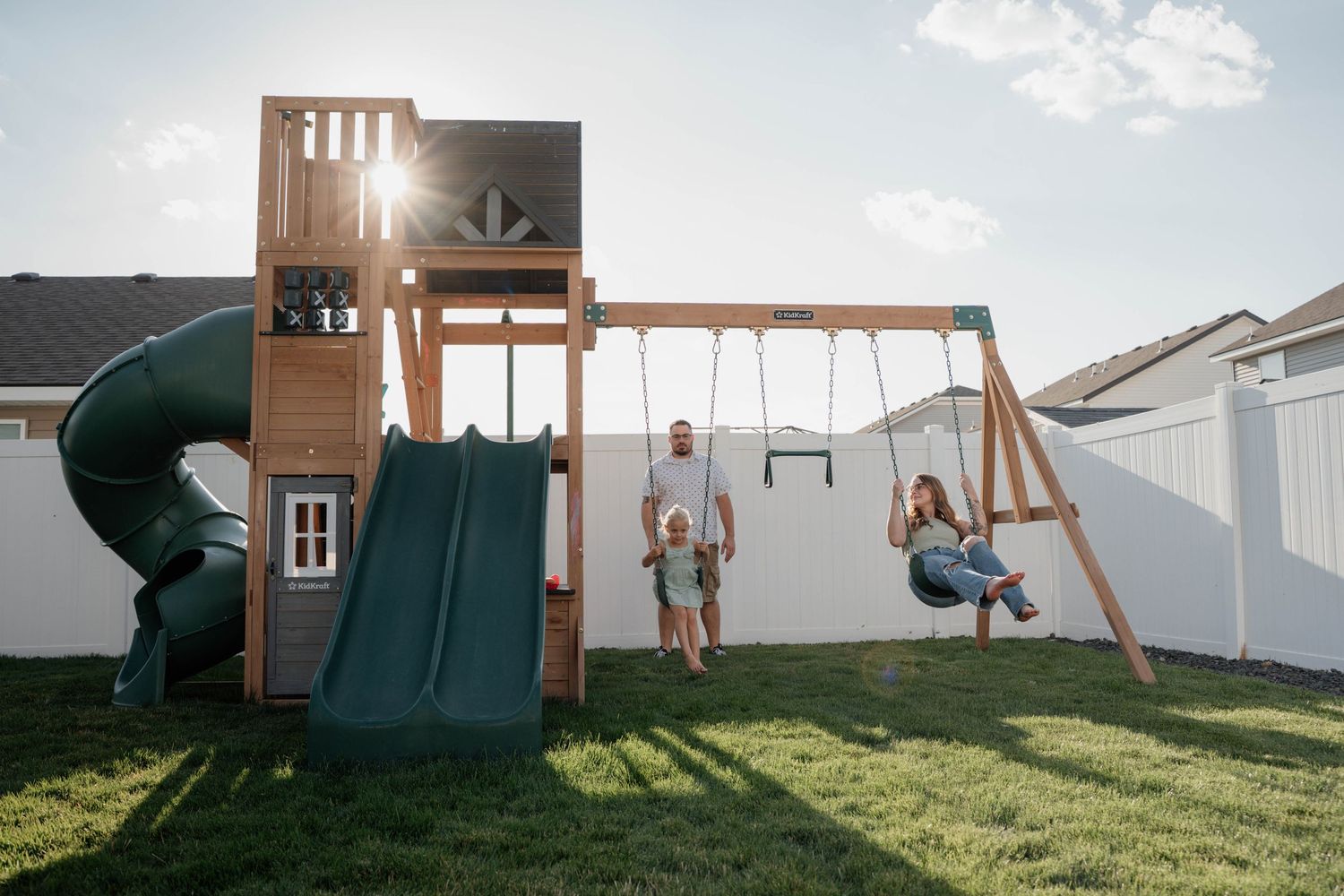 Family enjoying a backyard wooden swing set with green slide, tube slide, and swings on a sunny day.