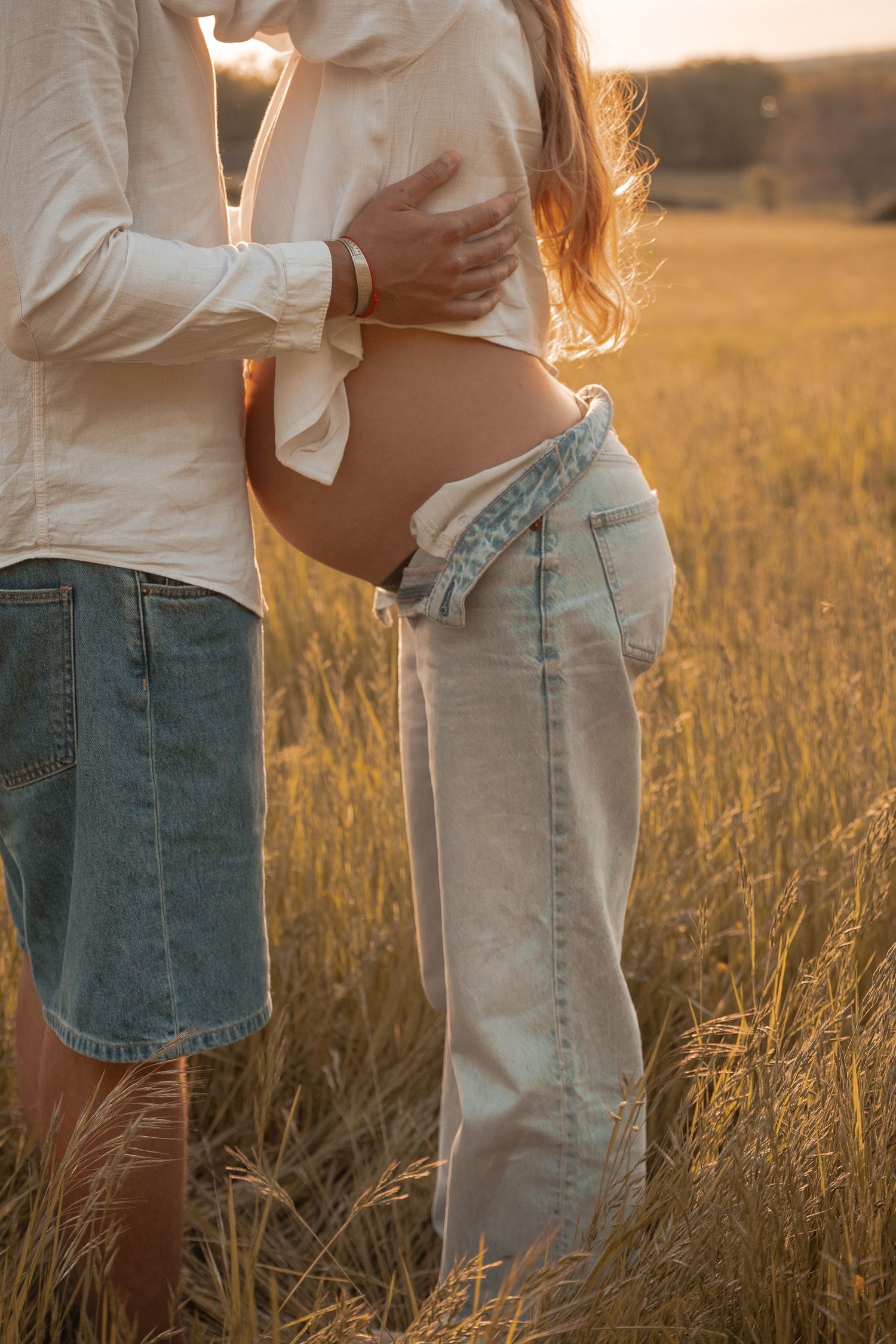 Plan moyen d'un homme enlaçant une femme enceinte dans un champ doré au coucher du soleil, photographie maternité lifestyle en Dordogne.