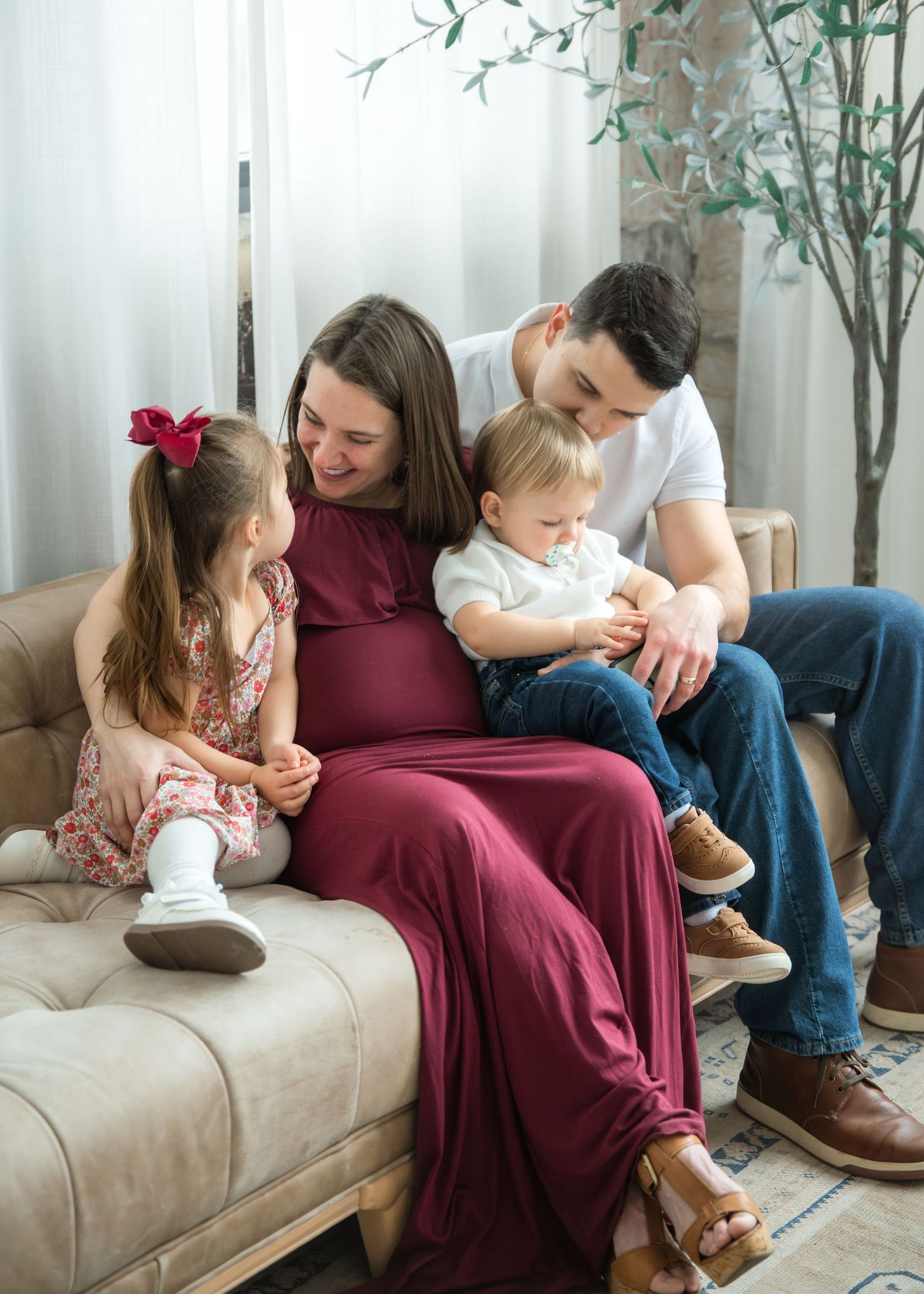 A pregnant woman in a burgundy dress sits on a beige sofa with her husband and two young children in a warm family portrait.