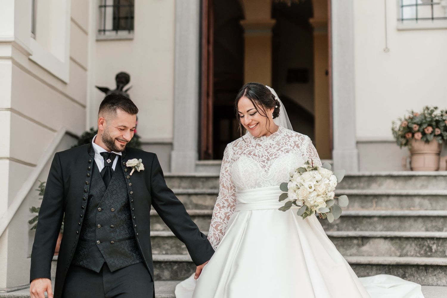 Bride in lace ball gown and groom in dark suit laughing on steps of elegant building.