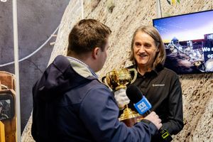 Man being interviewed while holding a gold Melbourne Cup trophy at an indoor event.