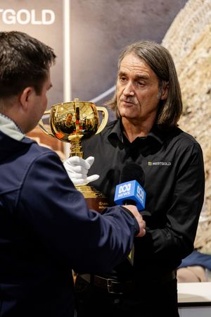 Man in Westgold shirt receiving the Melbourne Cup trophy from a white-gloved handler during ABC interview.