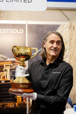 Man in black Westgold shirt holds gold Lexus Melbourne Cup trophy wearing white gloves at mining exhibition booth.