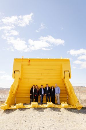 Group of five people posing in front of a large yellow mining bucket at an open-pit mine site.