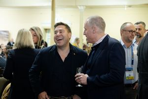 Two men laughing together while holding drinks at a crowded indoor social gathering event.