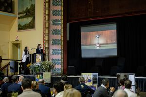 Two presenters at podium address seated audience in ornate hall with large projection screen displaying image.