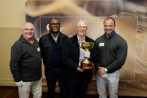 Four men posing together holding a gold trophy cup at a formal indoor event.