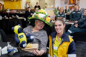 Woman in decorative floral hat holds gift box with staff member at Melbourne Cup celebration event.