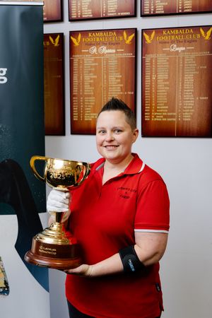 Woman in red shirt proudly holds gold Melbourne Cup trophy at Kambalda Football Club.