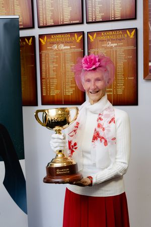 Elderly woman in pink fascinator holding the Lexus Melbourne Cup trophy at Kambalda Football Club.
