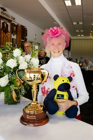 Elderly woman in pink fascinator smiling beside the Lexus Melbourne Cup trophy and a Westgold teddy bear.