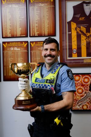 Police officer smiling and holding a gold Melbourne Cup trophy while wearing uniform in a football club room.