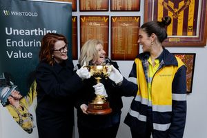 Three people smiling and holding a gold trophy cup near a Westgold mining company banner indoors.