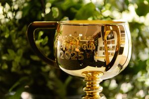 A shiny gold trophy cup engraved with the year 2025, displayed against a lush green leafy background.