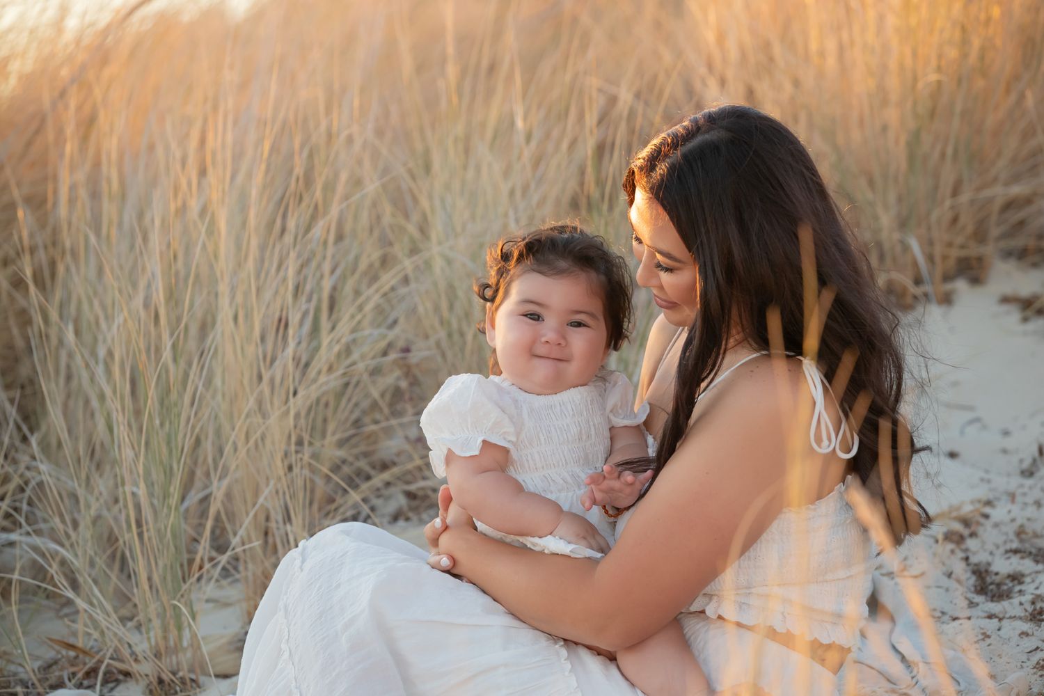 Mother in white dress cuddling smiling baby girl while sitting in sandy beach dunes at sunset.
