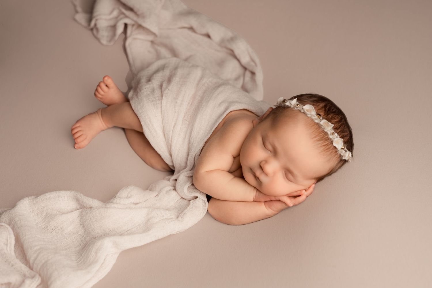 Sleeping newborn baby girl wrapped in soft white cloth with floral headband on neutral beige background.