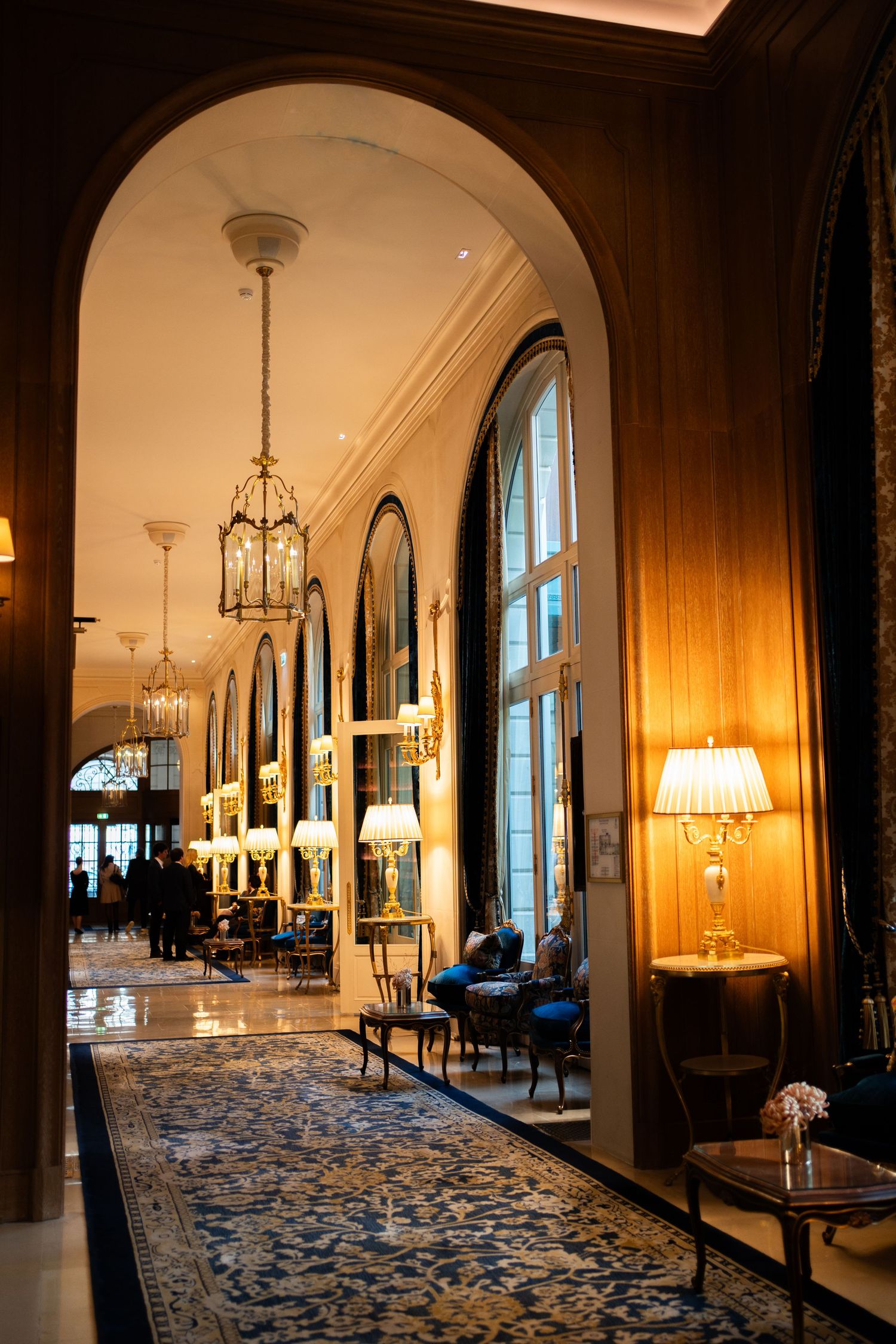 Elegant hotel corridor with arched ceilings, ornate chandeliers, tall windows, and a patterned carpet in warm golden lighting.