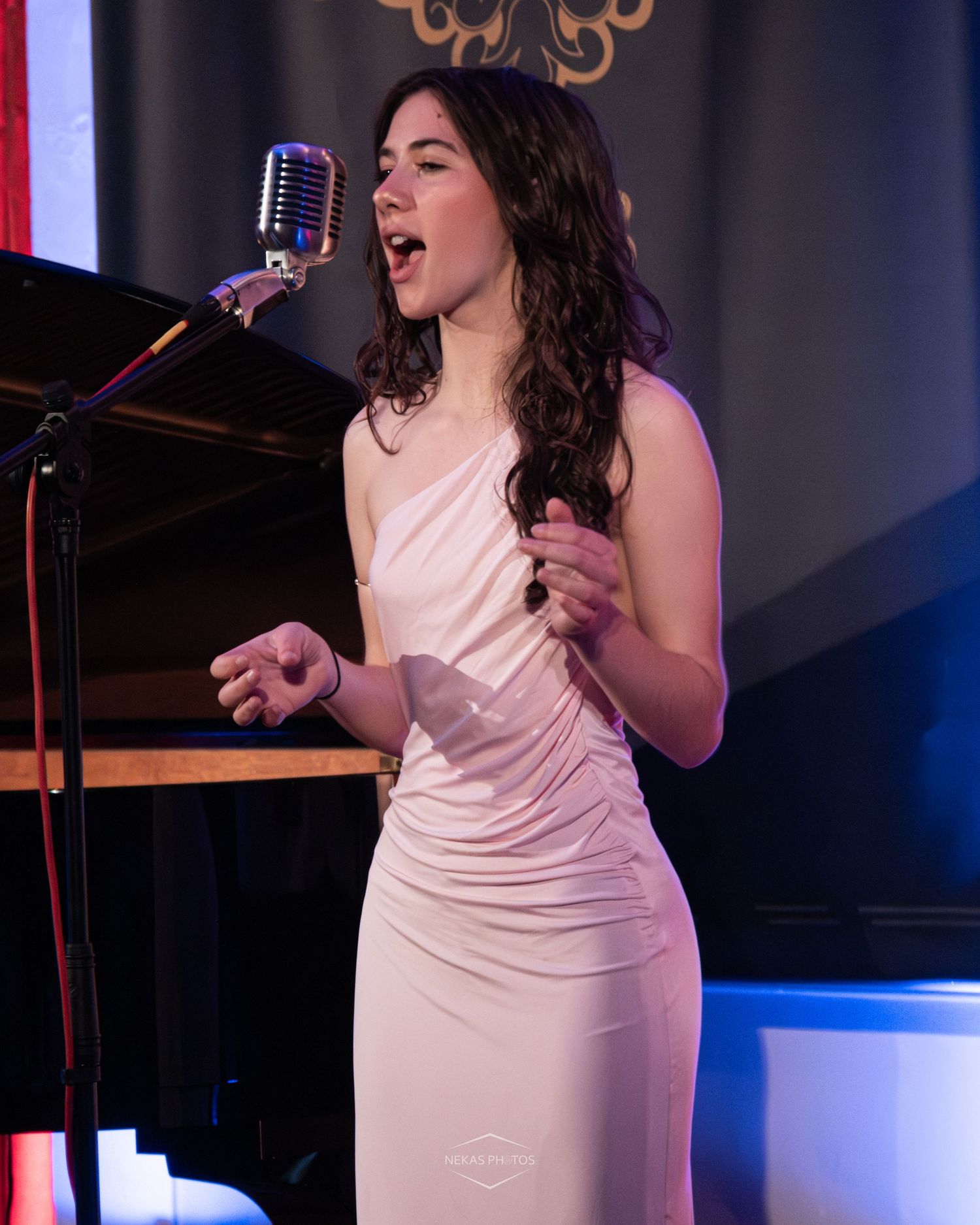 Young woman in white dress singing passionately into vintage microphone on stage at elegant venue.