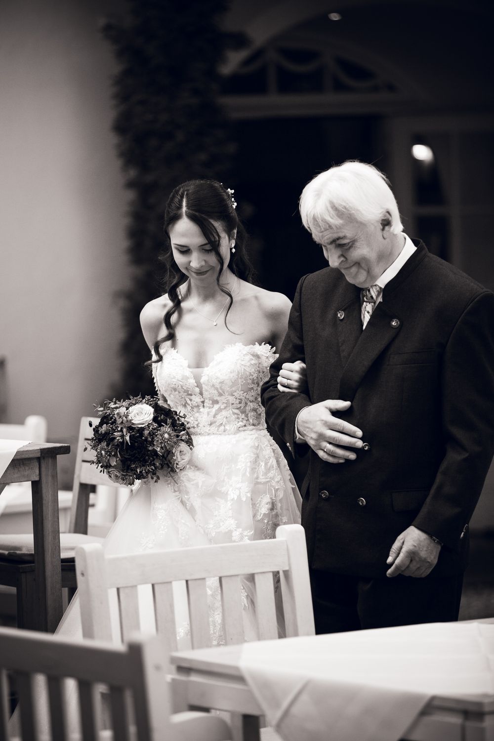 Bride in lace gown holding bouquet walks arm-in-arm with older man in black and white wedding photo.