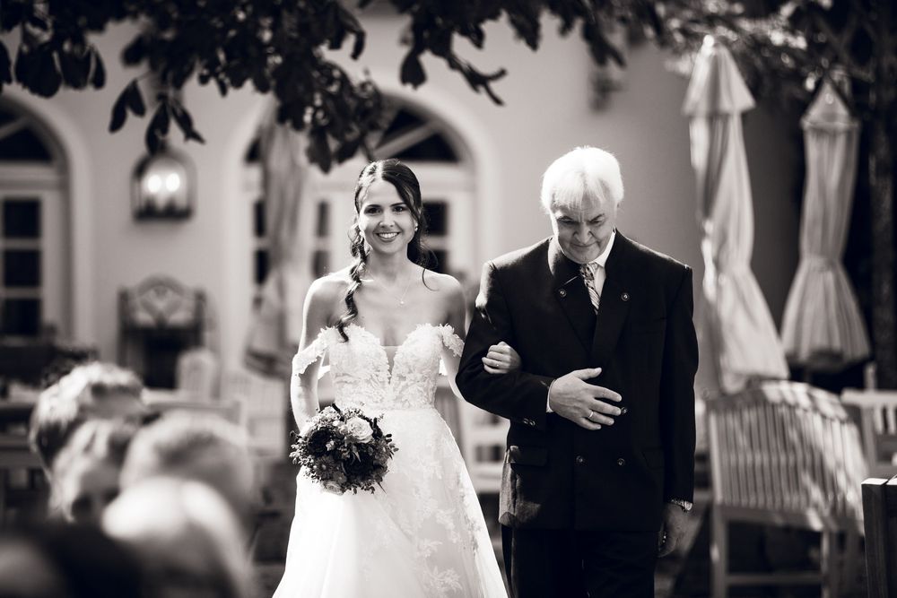 Smiling bride in white gown walks down outdoor aisle with elderly escort at elegant wedding venue.