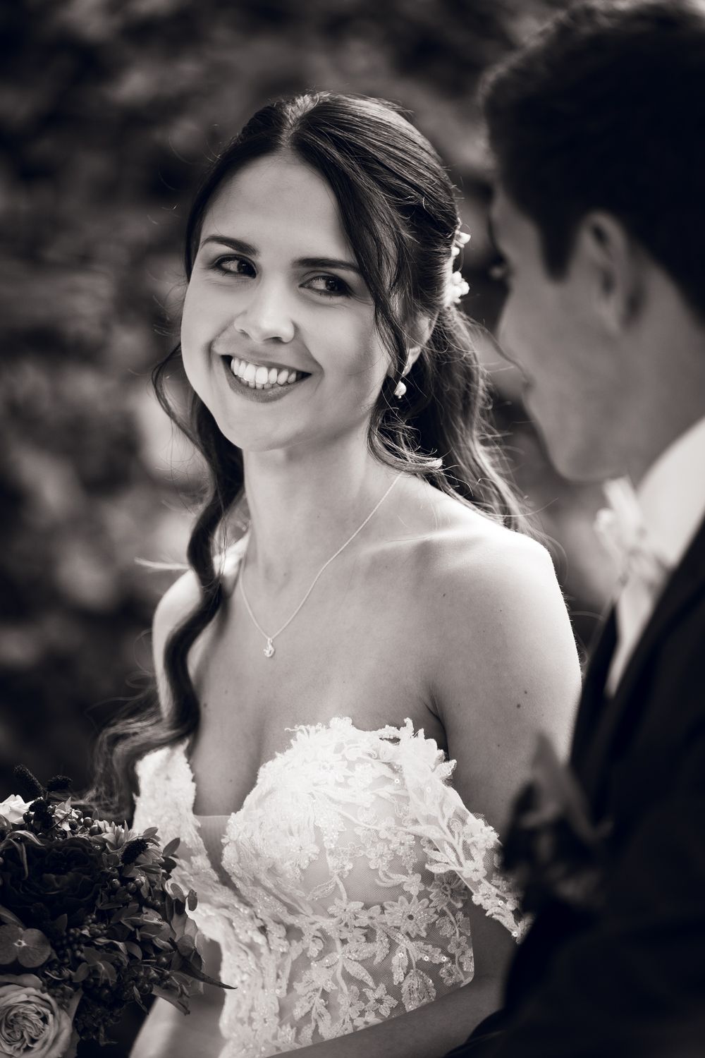 Smiling bride in lace off-shoulder gown holds bouquet while gazing at groom in black and white wedding photo.