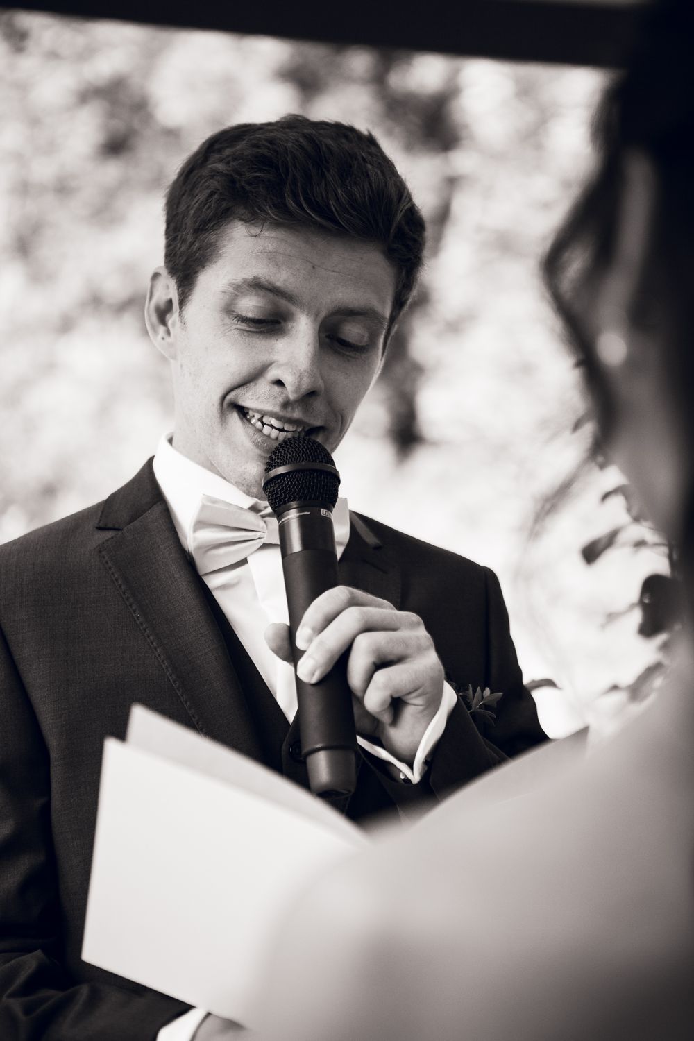 Groom in black tuxedo and bow tie reads wedding vows into microphone at outdoor ceremony.