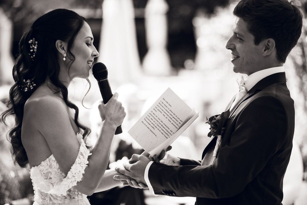 Bride reads heartfelt vows into microphone while groom smiles during outdoor wedding ceremony.
