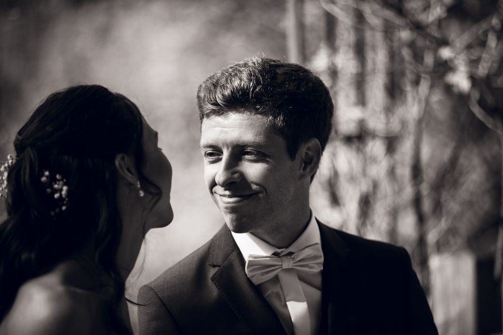 Black and white wedding photo of smiling groom in bow tie gazing at bride outdoors.