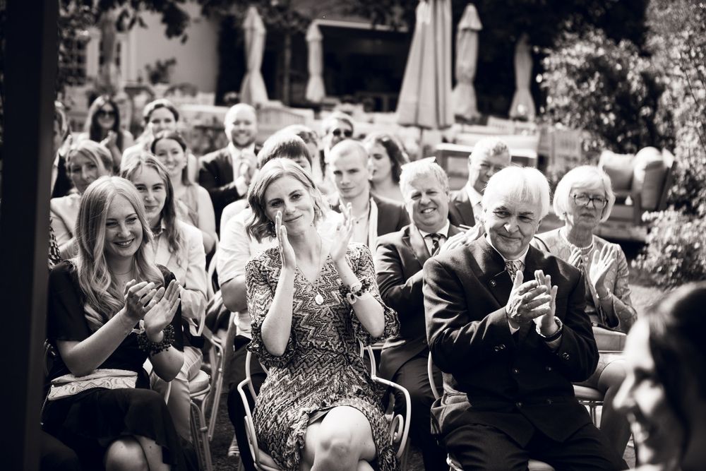 Black and white photo of wedding guests clapping and smiling during an outdoor ceremony.